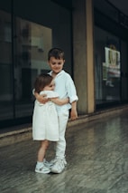 Two children are standing indoors, embracing each other warmly. Both are dressed in light-colored clothing, with one child wearing a white dress and sneakers, and the other child in a white shirt and pants. They are in a hallway with a polished marble floor and a glass window behind them.