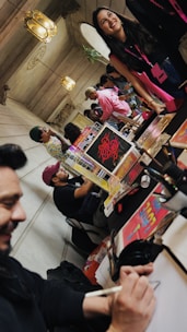 A lively indoor setting with people gathered around tables displaying colorful posters and materials. One person is drawing or writing, while others are engaged in conversation. Decorative lamps hang from the ceiling, and the atmosphere appears relaxed and social.
