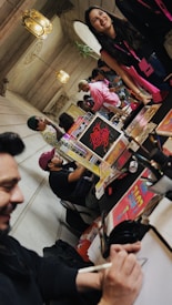 A lively indoor setting with people gathered around tables displaying colorful posters and materials. One person is drawing or writing, while others are engaged in conversation. Decorative lamps hang from the ceiling, and the atmosphere appears relaxed and social.