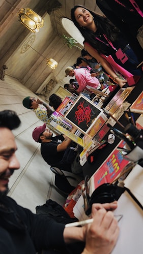 A lively indoor setting with people gathered around tables displaying colorful posters and materials. One person is drawing or writing, while others are engaged in conversation. Decorative lamps hang from the ceiling, and the atmosphere appears relaxed and social.