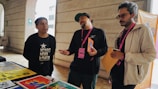 Three individuals are standing near a table displaying colorful posters and papers. One person is leaning slightly backward, appearing to speak or express an idea, while another listens intently with his arms holding papers. The environment suggests an informal meeting or discussion.