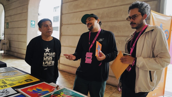 Three individuals are standing near a table displaying colorful posters and papers. One person is leaning slightly backward, appearing to speak or express an idea, while another listens intently with his arms holding papers. The environment suggests an informal meeting or discussion.