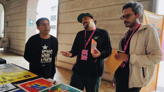 Three individuals are standing near a table displaying colorful posters and papers. One person is leaning slightly backward, appearing to speak or express an idea, while another listens intently with his arms holding papers. The environment suggests an informal meeting or discussion.