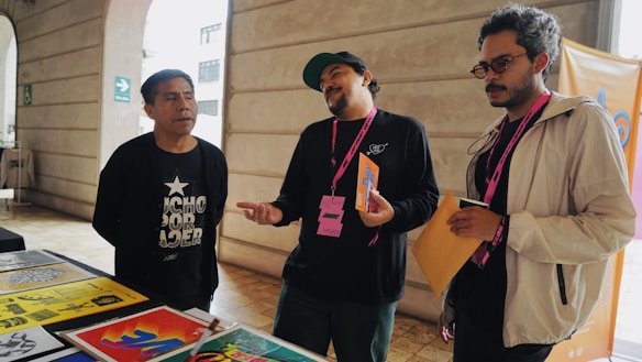 Three individuals are standing near a table displaying colorful posters and papers. One person is leaning slightly backward, appearing to speak or express an idea, while another listens intently with his arms holding papers. The environment suggests an informal meeting or discussion.
