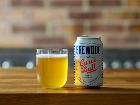 A can of BrewDog Curve Ball West Coast IPA is placed next to a glass filled with light amber beer on a wooden surface. The background features a blurred brick wall, adding a rustic feel to the scene.