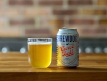 A can of BrewDog Curve Ball West Coast IPA is placed next to a glass filled with light amber beer on a wooden surface. The background features a blurred brick wall, adding a rustic feel to the scene.