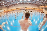 A large group of people dressed in white martial arts uniforms are practicing techniques in a gymnasium with a blue floor. The space has a high ceiling with metal beams and bright lights. There are spectators sitting on an upper level gallery overlooking the group.