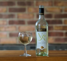 A bottle of Hardy's VR Pinot Grigio 2022 is placed next to a filled wine glass on a wooden surface. The background consists of a blurred brick wall, giving a rustic and cozy atmosphere.