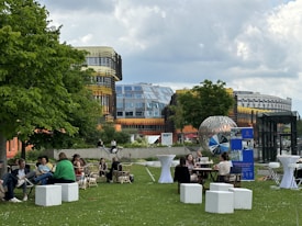A group of people are gathered outdoors on a grassy lawn, seated around tables and on benches. The setting is in front of modern and colorful architectural buildings with glass windows and unique designs. There is a large decorative metallic sphere displayed prominently, and some promotional banners or signs are visible. The scene suggests a casual social or professional gathering under partially cloudy skies.