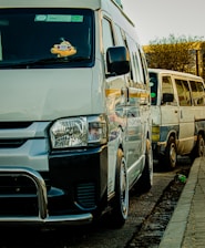 Two vans are parked along a street. The van in the foreground is white with a black bumper and a stuffed toy hanging from the rearview mirror. The second van, situated behind the first one, is an older model with visible wear.