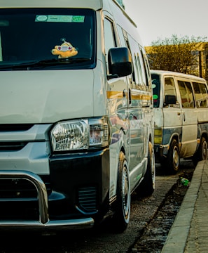 Two vans are parked along a street. The van in the foreground is white with a black bumper and a stuffed toy hanging from the rearview mirror. The second van, situated behind the first one, is an older model with visible wear.