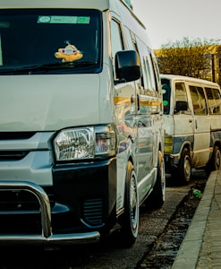 Two vans are parked along a street. The van in the foreground is white with a black bumper and a stuffed toy hanging from the rearview mirror. The second van, situated behind the first one, is an older model with visible wear.
