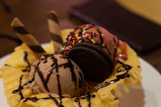 A delicious dessert featuring scoops of chocolate and strawberry ice cream sitting in a waffle bowl. The ice cream is topped with colorful sprinkles, a chocolate cookie, and two wafer sticks. Chocolate syrup is drizzled over the entire creation.