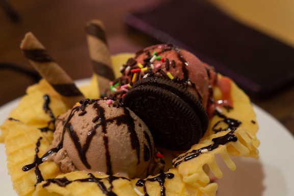 A delicious dessert featuring scoops of chocolate and strawberry ice cream sitting in a waffle bowl. The ice cream is topped with colorful sprinkles, a chocolate cookie, and two wafer sticks. Chocolate syrup is drizzled over the entire creation.