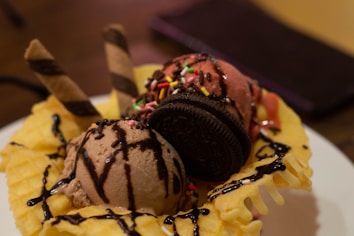 A delicious dessert featuring scoops of chocolate and strawberry ice cream sitting in a waffle bowl. The ice cream is topped with colorful sprinkles, a chocolate cookie, and two wafer sticks. Chocolate syrup is drizzled over the entire creation.