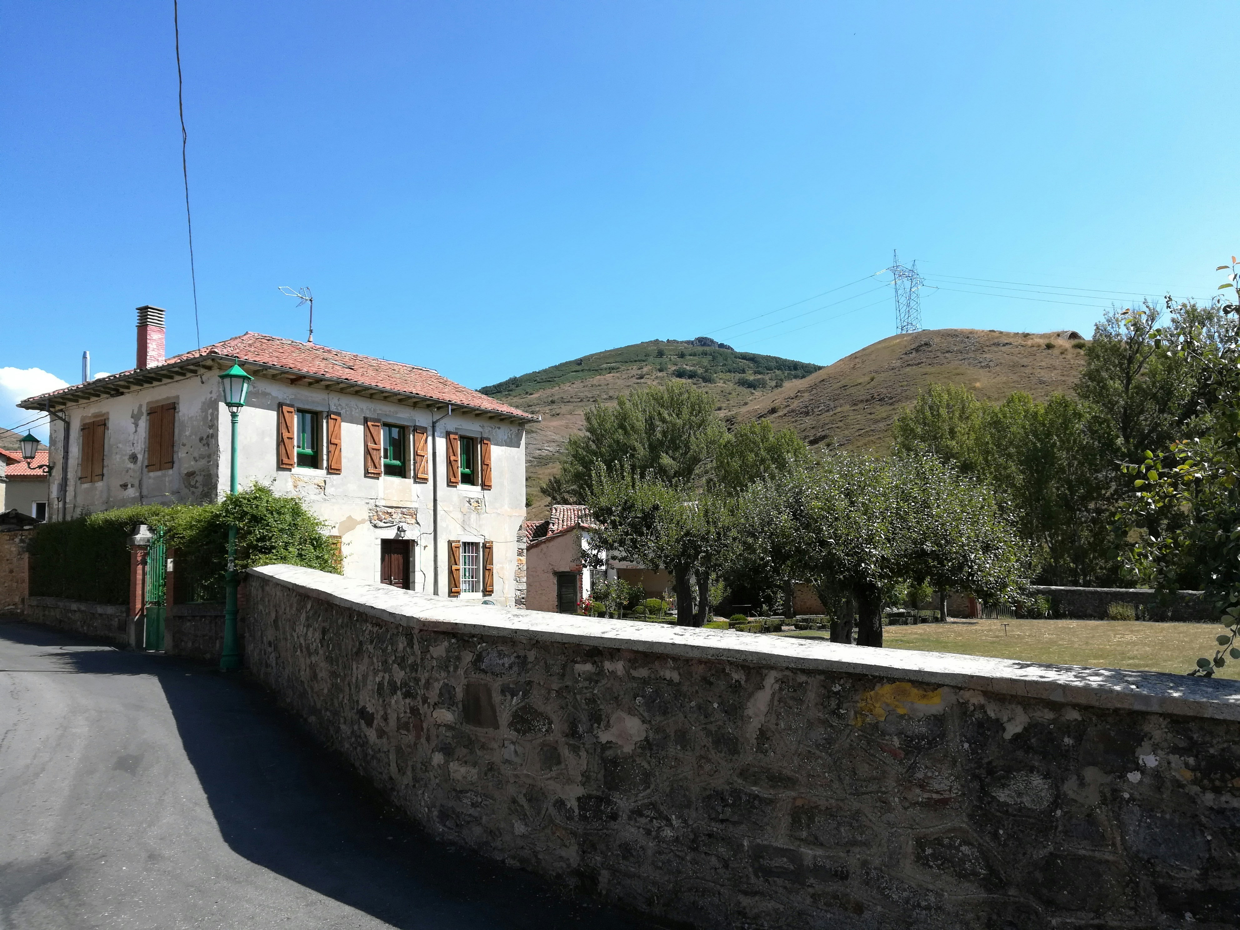 A rustic two-story house with a red-tiled roof and brown window shutters stands against a backdrop of green hills. The property is surrounded by a stone wall, and there are trees and a small garden area in the front yard. An electricity pylon and cables are visible in the distance, and a clear blue sky adds a serene feel to the scene.