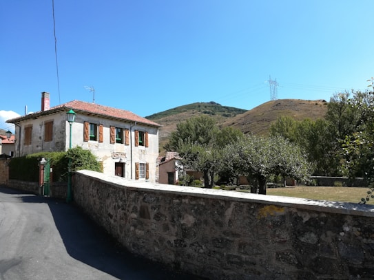 A rustic two-story house with a red-tiled roof and brown window shutters stands against a backdrop of green hills. The property is surrounded by a stone wall, and there are trees and a small garden area in the front yard. An electricity pylon and cables are visible in the distance, and a clear blue sky adds a serene feel to the scene.