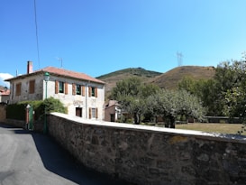 A rustic two-story house with a red-tiled roof and brown window shutters stands against a backdrop of green hills. The property is surrounded by a stone wall, and there are trees and a small garden area in the front yard. An electricity pylon and cables are visible in the distance, and a clear blue sky adds a serene feel to the scene.