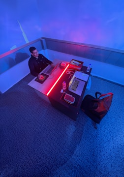A person sits at a desk in a modern office environment, working on a laptop. The lighting is ambient with shades of blue and red creating a futuristic atmosphere. The desk is neatly organized with another laptop, a notepad, water bottles, and some wires. The carpeted floor adds a feeling of coziness to the workspace.