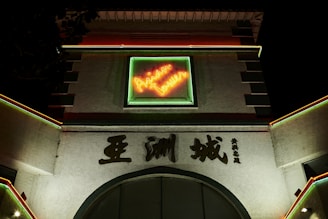A building facade with a prominent neon sign that says 'Asian Tower' in bright orange and green colors. The structure has architectural elements like archways and is adorned with Asian characters. It is illuminated with colorful lights at night, creating a vibrant and lively atmosphere.