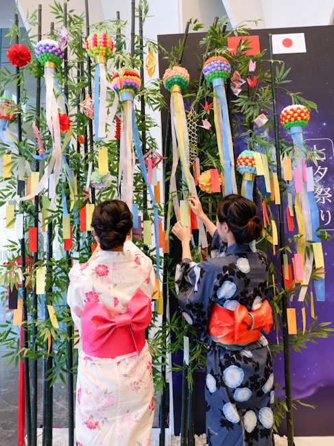 Two women dressed in traditional Japanese attire, known as yukata, are placing colorful paper strips on tall bamboo branches. The display features vibrant pom-poms, ribbons, and decorations consistent with a festival atmosphere. Japanese flags and seasonal motifs complement the decorative scene.