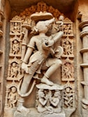 An intricately carved stone relief depicting a mythological figure with a human body and an animal head, surrounded by smaller figures. The main figure is adorned with detailed jewelry and traditional attire, striking a dynamic pose. Surrounding figures and ornate elements suggest a narrative or cultural significance.