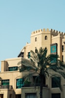 A beige building with traditional Middle Eastern architectural elements, including arched windows and decorative parapets. A tall palm tree stands prominently in the foreground, and the sky is clear and blue.