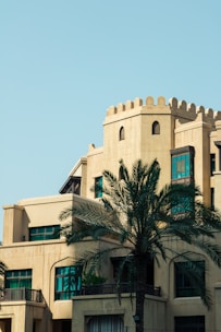 A beige building with traditional Middle Eastern architectural elements, including arched windows and decorative parapets. A tall palm tree stands prominently in the foreground, and the sky is clear and blue.