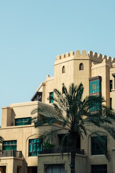 A beige building with traditional Middle Eastern architectural elements, including arched windows and decorative parapets. A tall palm tree stands prominently in the foreground, and the sky is clear and blue.