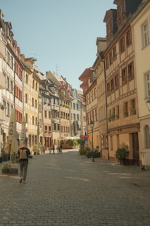 A picturesque, cobblestone street lined with colorful, traditional European buildings featuring wooden frames and flower boxes. Several people are leisurely walking, and bicycles and scooters are visible along the sidewalk. The street has a quaint and historical atmosphere with outdoor caf&eacute; umbrellas in the distance.