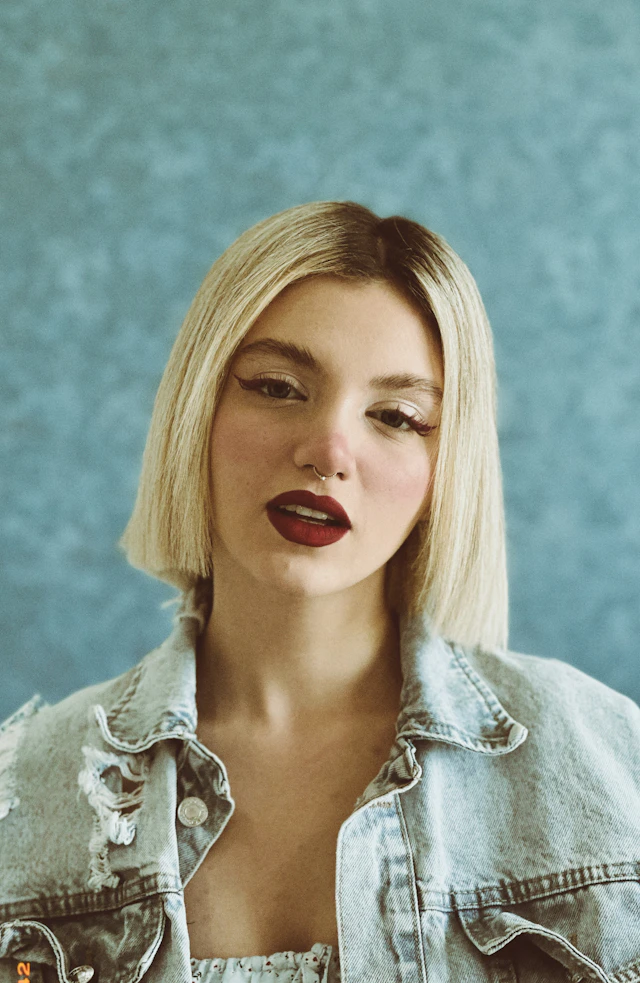 Margaux in a faded denim jacket against a blue wall, looking into the camera.