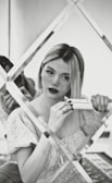 A woman with blonde hair is using a hair straightener while looking into a decorative mirror with a grid design. She is wearing a floral-patterned blouse and has a focused expression. The image is in black and white, giving it a classic and vintage feel.