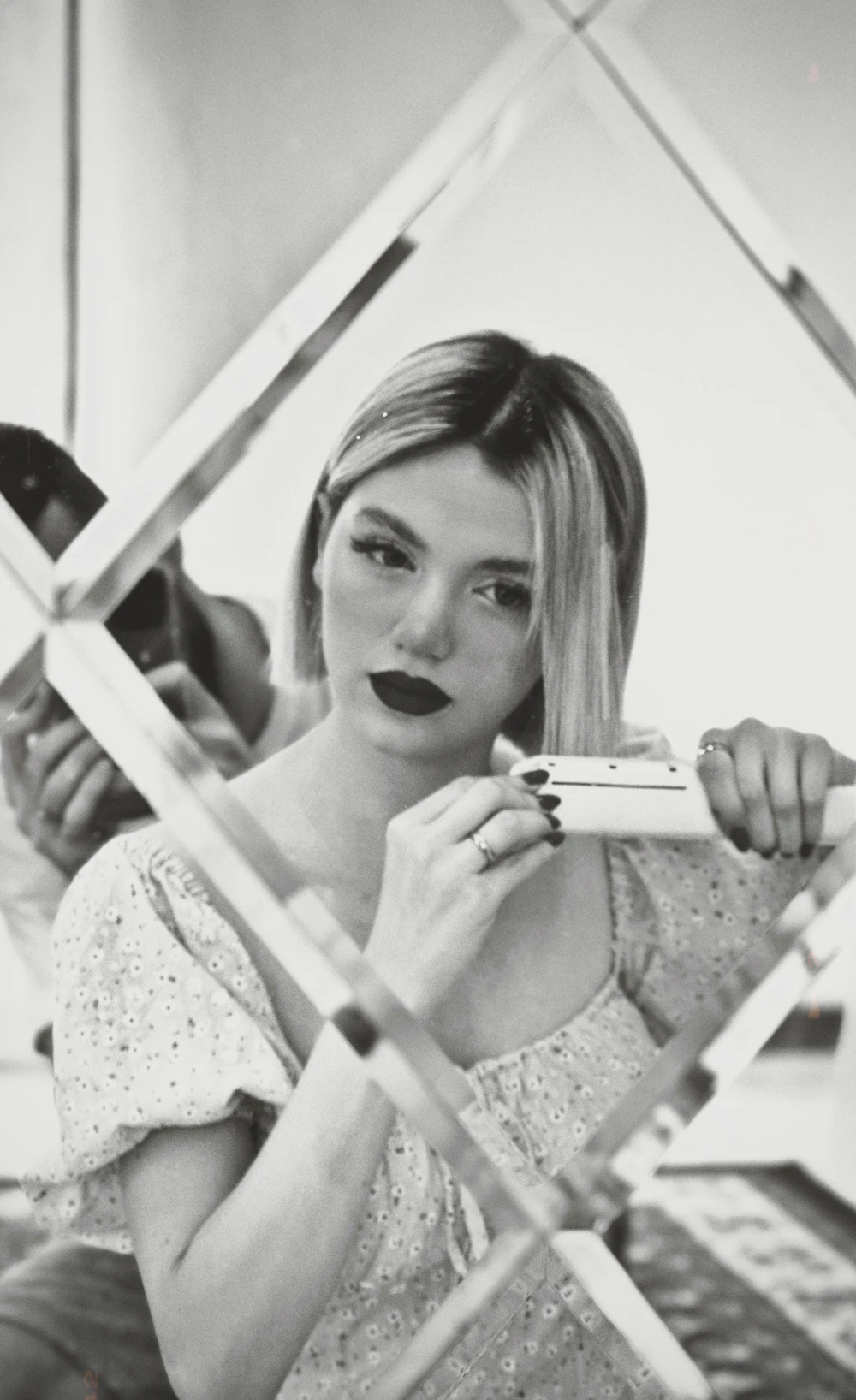 Black and white photo of Margaux reflected in broken mirror panels, holding a compact.