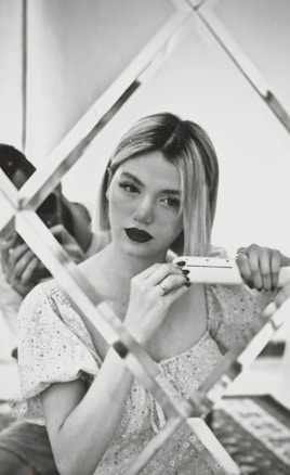 A woman with blonde hair is using a hair straightener while looking into a decorative mirror with a grid design. She is wearing a floral-patterned blouse and has a focused expression. The image is in black and white, giving it a classic and vintage feel.