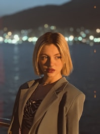 A person with short blonde hair is wearing a suit jacket over a patterned shirt. The background features a body of water with bokeh lights from a distant shoreline, suggesting an evening scene.