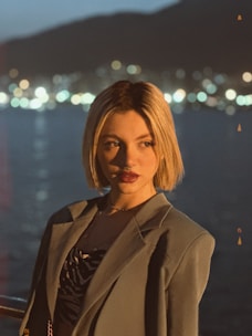 A person with short blonde hair is wearing a suit jacket over a patterned shirt. The background features a body of water with bokeh lights from a distant shoreline, suggesting an evening scene.