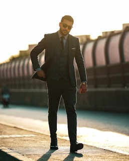 A man in a dark suit walks confidently on a sidewalk. He is wearing sunglasses and a wristwatch, with his suit jacket partially open. The background is blurred, with a structure and bright sunlight casting long shadows.