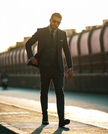 A man in a dark suit walks confidently on a sidewalk. He is wearing sunglasses and a wristwatch, with his suit jacket partially open. The background is blurred, with a structure and bright sunlight casting long shadows.