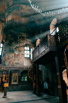 The interior of an ornate, historical church with high ceilings and detailed frescoes decorating the walls. Sunlight streams through the windows, creating luminous beams that illuminate the space. An intricately carved wooden balcony runs along the upper part of the wall, and a spiral staircase provides access to it. Candles are lit near a religious icon, and a person stands in silent contemplation.
