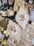 A collection of vintage sewing and crafting items is arranged on a surface, including two framed floral embroidery pieces, antique books, scissors, spools of pink thread, sheet music, and lace. The embroidery features delicate pink roses with green leaves, housed in ornate, off-white frames. The overall aesthetic combines elements of vintage charm and artistic craftsmanship.