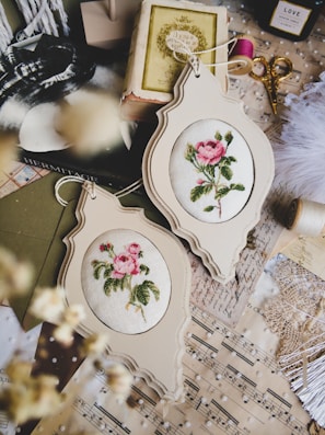 A collection of vintage sewing and crafting items is arranged on a surface, including two framed floral embroidery pieces, antique books, scissors, spools of pink thread, sheet music, and lace. The embroidery features delicate pink roses with green leaves, housed in ornate, off-white frames. The overall aesthetic combines elements of vintage charm and artistic craftsmanship.