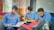 Three young men, dressed in matching blue shirts and red lanyards, are sitting closely at a table. They appear engaged in a discussion, focused on a notebook that one of them is holding open. The setting seems to be a classroom or study area with windows in the background and a bag on the table.