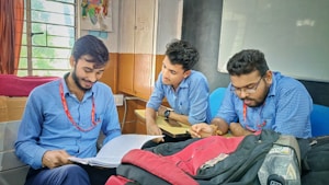 Three young men, dressed in matching blue shirts and red lanyards, are sitting closely at a table. They appear engaged in a discussion, focused on a notebook that one of them is holding open. The setting seems to be a classroom or study area with windows in the background and a bag on the table.