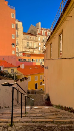 A narrow cobblestone street lined with colorful buildings featuring vibrant red, yellow, and pastel hues. The street slopes downhill and is framed by a railing on the left. The sky is clear blue, and sunlight casts warm tones on the architecture.