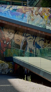 A vibrant and colorful mural depicting various cartoon and comic book characters is painted on the walls beneath a modern pedestrian bridge. The artwork features bold lines and exaggerated expressions, set against the backdrop of an urban environment with glass and metal architecture.