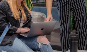 One person sits on a sofa using a laptop while another stands nearby, pointing at the screen. The seated individual wears a black jacket and jeans with a ripped knee, and the standing person is in pinstriped pants. A tumbler and a potted plant are visible in the surroundings.