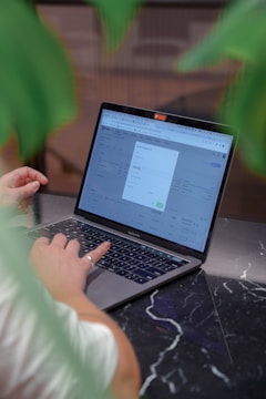 A person is using a MacBook Pro laptop, which is placed on a dark marble table. The screen displays a webpage with an open dialogue box, suggesting an online activity or transaction. Green leaves partially frame the image, indicating a natural or relaxed setting.