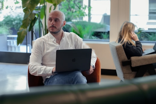 A man with a shaved head is seated on an orange chair indoors, using a laptop. He is wearing a white shirt and looking to the side. In the background, a woman with long hair is sitting on a cushioned seat, talking on a phone and also using a laptop. The setting appears to be a modern office or co-working space with large windows and greenery outside.