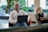 A man with a shaved head is seated on an orange chair indoors, using a laptop. He is wearing a white shirt and looking to the side. In the background, a woman with long hair is sitting on a cushioned seat, talking on a phone and also using a laptop. The setting appears to be a modern office or co-working space with large windows and greenery outside.