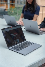 Three people are seated at a long table, each using a laptop. The laptops are Apple MacBooks. One of the laptops has a screen displaying a website with the text 'Where projects run smoothly.' The setting appears to be a modern, well-lit indoor space with greenery visible in the background.